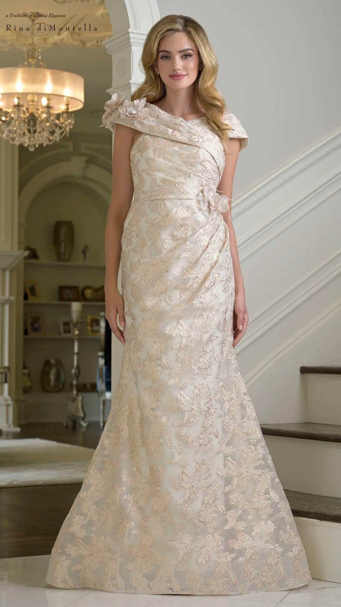 A woman stands indoors in a Rina Di Montella RD3428 long formal dress in champagne, with cap sleeves, subtle draping, and a graceful mermaid skirt, in front of a staircase and chandelier.